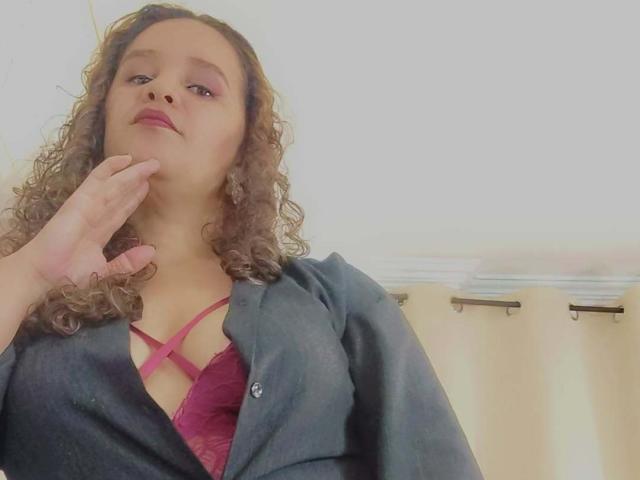 VittoriaI with curly brown hair, wearing a pink strappy bra under a dark blue shirt, touches her chin with her right hand, looking slightly up.