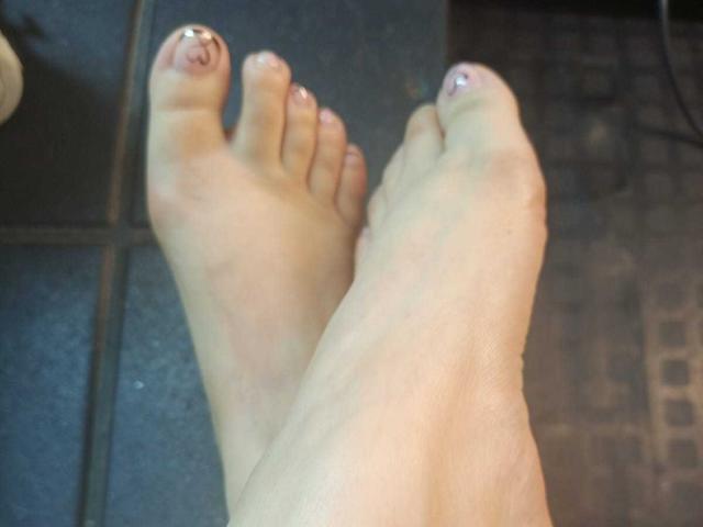 BelaDeimons' feet with shiny, glittery toenails, showing the big toes and the rest of the toes crossed, on a dark tiled floor.