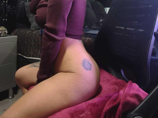 AmelieBlake, wearing a red ribbed long-sleeve top, is kneeling on a red towel, exposing her bare ass with a sun tattoo on her right buttock, in a chair.