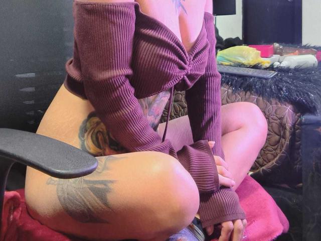 AmelieBlake sits cross-legged on a chair, wearing a ribbed, purple, off-shoulder top, exposing her tits and a large tattoo on her right thigh. Her ass is visible.
