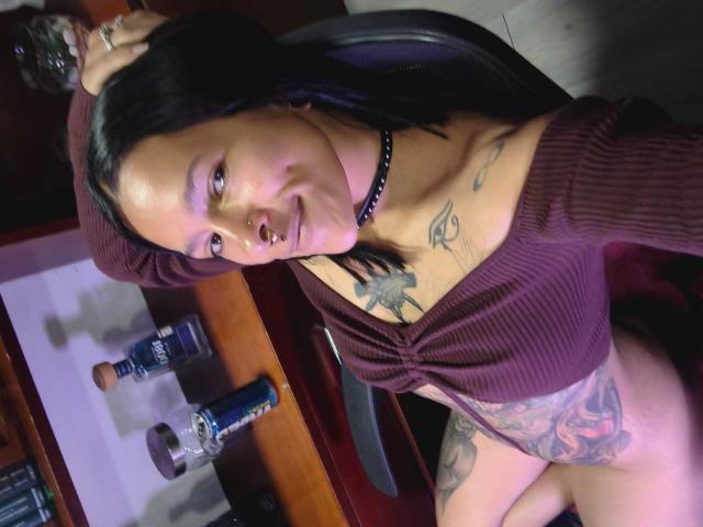 AmelieBlake with black hair, nose ring, choker, tattoos, and small tits, wearing a maroon top, sitting, smiling, and showing her bare pussy in a slightly tilted photo.