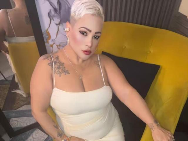 Gianella Cox, con cabello rubio corto, vestido blanco, busto grande, tatuajes, lápiz labial rojo, sentada en una silla amarilla, mirando hacia arriba, con un brazo apoyado en la silla, reflejo en un espejo detrás..