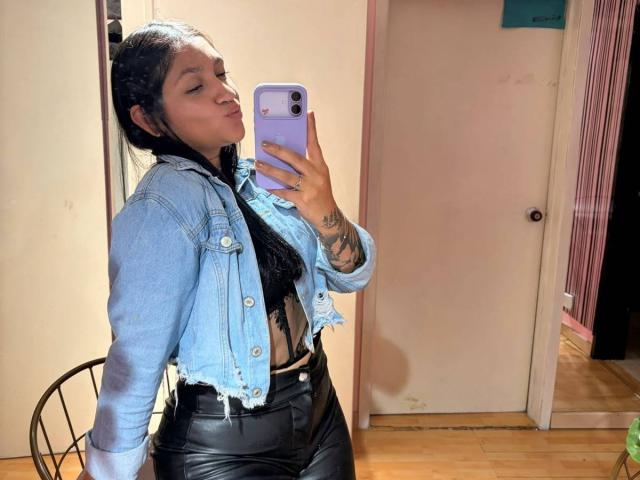 MiaaVixen with puffy lips, wearing a blue denim jacket, black lace top, and black leather pants, holding a purple phone, showing tattoos on her hand.