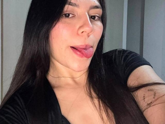 SophiaMarx, con el pelo largo y negro, sacando la lengua de color rosa, vistiendo una camisa negra, mostrando un pequeño tatuaje en su brazo derecho y un collar delgado..