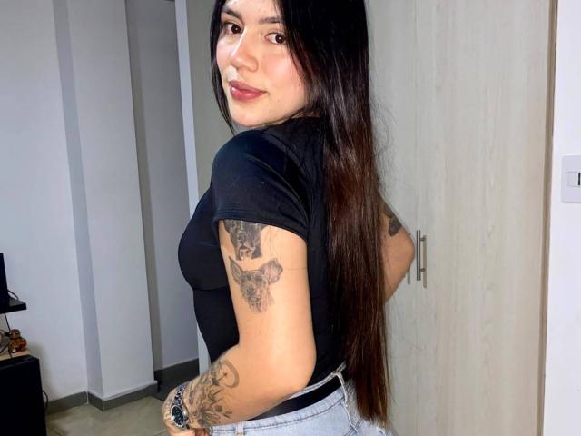 SophiaMarx with long black hair, black t-shirt, tattoos on arms, light grey pants, smiling at camera, standing in front of wooden door, showing her small, perky ass.