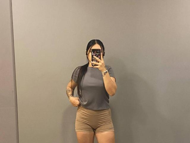SophiaMarx, con lunghi capelli neri, indossa una maglietta grigia e dei pantaloncini beige, è in piedi di fronte a un muro grigio, tiene in mano un telefono e mostra dei tatuaggi sul braccio destro..