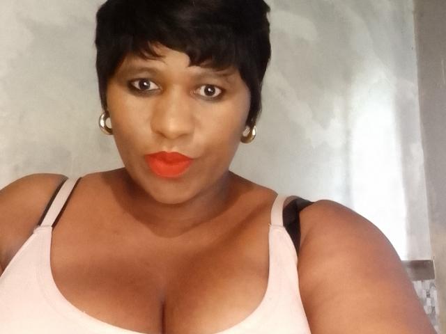 TracySmith with short black hair, red lipstick, hoop earrings, and a white tank top showing her big tits. She's looking at the camera with a slight pout.