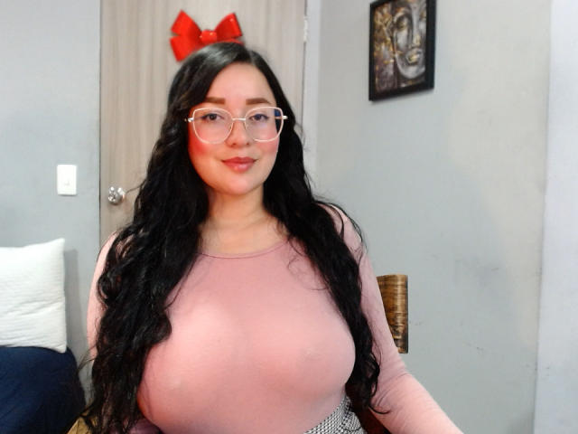 JaneAndPaull, con senos grandes, vistiendo una blusa rosa, gafas y un lazo rojo, tiene el pelo largo y negro, y una sonrisa encantadora en un dormitorio..