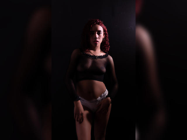 ValeryMartinez stands in dark lighting, wearing a sheer black top showing her small tits, and white lace panties, with red curly hair, and a serious expression.
