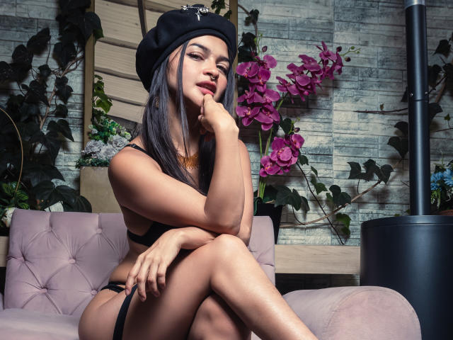LucyySky with long black hair, wearing a black beret and black lingerie, sits on a pink couch, finger on her red lips, crossed legs, purple orchids in background.