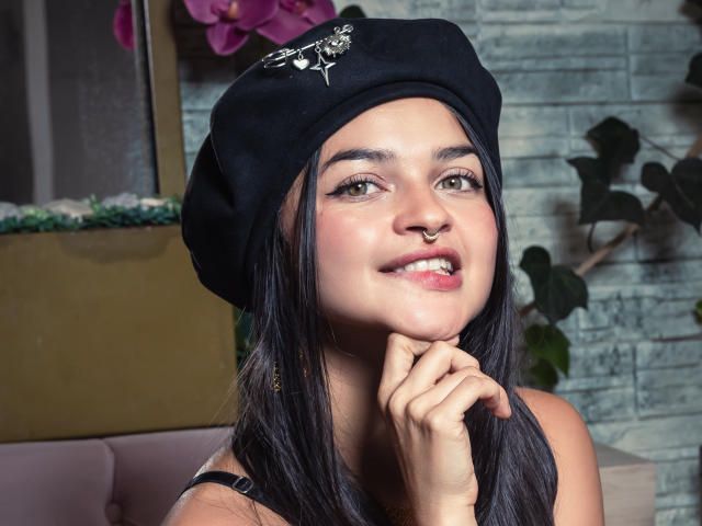 LucyySky with long black hair, black beret, nose ring, and smile, touching chin, showing off her pretty face and subtle makeup, sitting with a relaxed, cute expression.