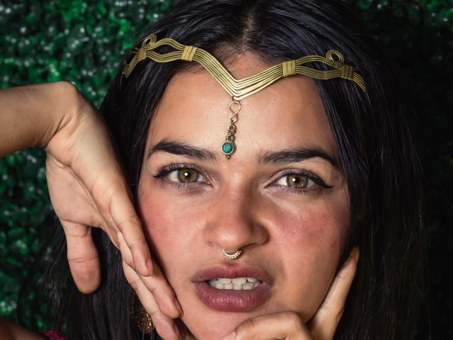 LucyySky's face with brown eyes, dark hair, gold headpiece, green gem, nose ring, and hands touching cheeks, showing slightly parted lips. She's wearing subtle makeup.