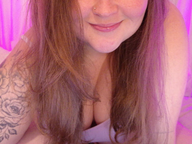 Lilysweetxx with long brown hair, smiling, nose pierced, showing her large boobs, tattooed arm, and pinkish-purple lighting. She's on a bed, looking down at the camera.