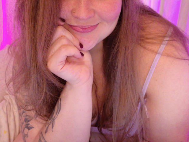Lilysweetxx with pinkish skin, brown hair, and a nose piercing, smiling and resting her chin on her hand, showing a tattoo and purple nail polish, in a pink-lit room.
