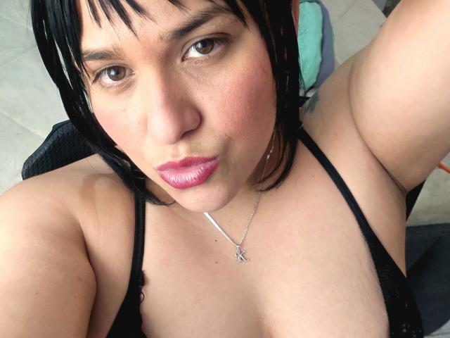 Viviana Thompson tem batom rosa, cabelo preto e um colar de prata com um pingente em forma de 'K'. Ela está usando um sutiã preto, mostrando seus seios, e tem uma expressão de lábios franzidos..