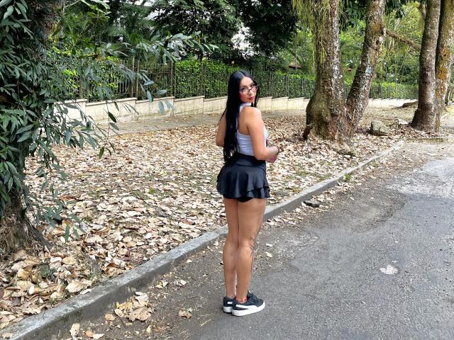 ChloeMoretti stands on a leafy path, looking back with glasses, black skirt, gray top, and black sneakers, showing her ass and curvy, tan-skinned body with long black hair.