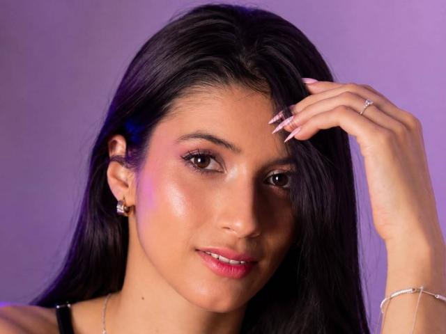 JasminKooper ha i capelli neri e lisci, gli occhi marroni, il rossetto rosa e la pelle luminosa. Sta toccando i capelli con una mano adornata da un anello, indossa orecchini e un braccialetto..