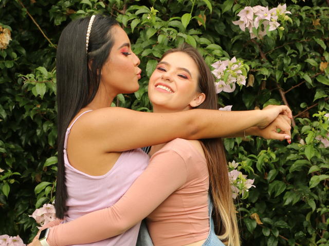 PaolaYKatty with long black hair, pink tank, and white headband kissing another PaolaYKatty with brown hair, pink top, and blue overalls, arms around each other's shoulders.