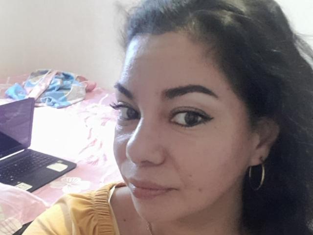 HypnoticBella with dark curly hair, hoop earrings, and thick eyeliner, looking at a laptop on a pink bed, wearing a yellow top, showing a slight pimple on her nose.
