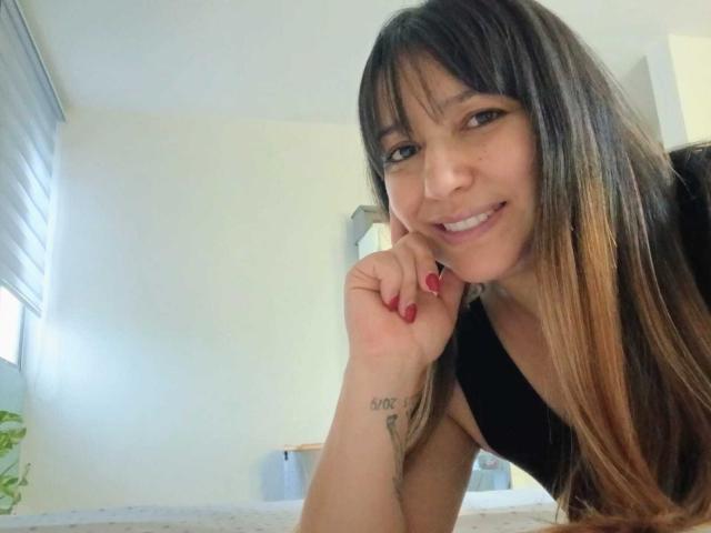 RoxanneBlair with long brown hair, bangs, and a black top, smiling at the camera, right hand with red nails and "SOO" tattoo, in a bright room.