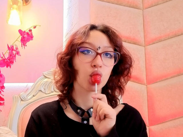 JinXkm with curly red hair, glasses, black choker, and black top, sucking a red lollipop, has a tattoo above her left eyebrow, and is looking at the camera.
