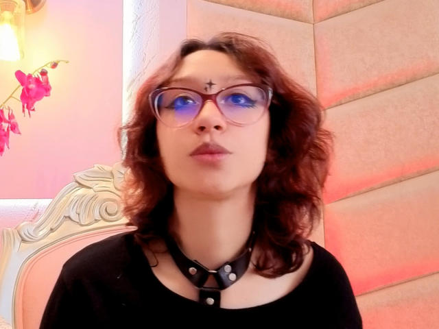 JinXkm with curly brown hair, red glasses, black choker, and cross forehead dot, wearing black top, looking up with slightly pouted lips, soft pink and orange lighting.