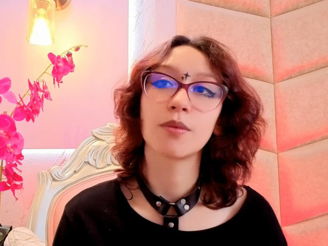 JinXkm has curly red hair, blue-tinted glasses, a black star on her forehead, and a black choker with silver studs, wearing a black shirt, with pink flowers in the background.