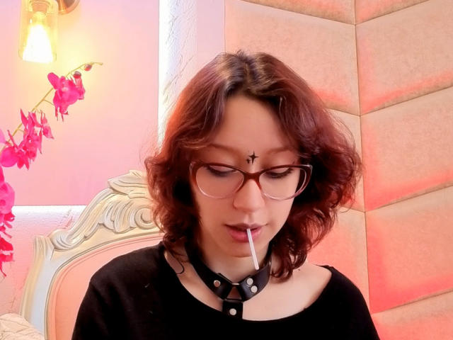 JinXkm with curly red-brown hair, glasses, black choker, sucking on a straw, black top, nose piercing, pink flowers, and warm light. Soft pink and beige wall.