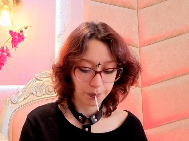 JinXkm with wavy brown hair, glasses, black choker with metal studs, black top, and a cigarette in mouth. She's got a small black cross on her forehead.