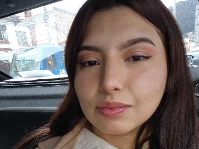 SofieMalory has puffy pinkish lips, brown eyes with winged eyeliner, and brown hair. She's wearing a beige coat, sitting in a car, with a blue bus outside.