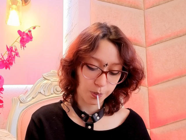 JinXkm with red curly hair, glasses, black shirt, and black collar with studs, smoking a cigarette, has a star tattoo on her forehead, pinkish lighting.