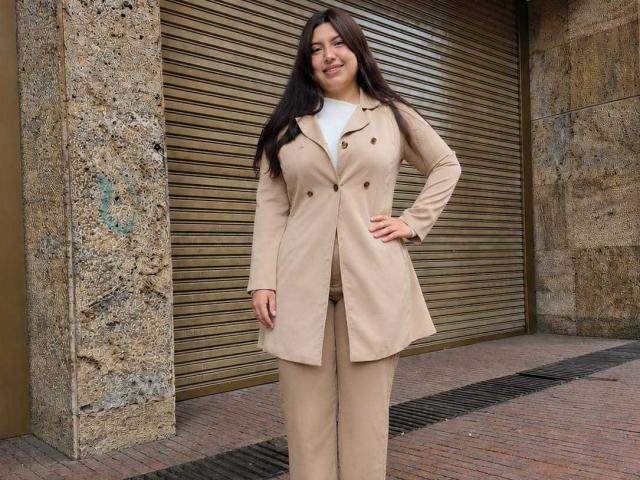 SofieMalory stands in front of a shut, metallic, brown roll-up door wearing a beige, double-buttoned, long coat with matching pants, white shirt, and long, dark brown hair.