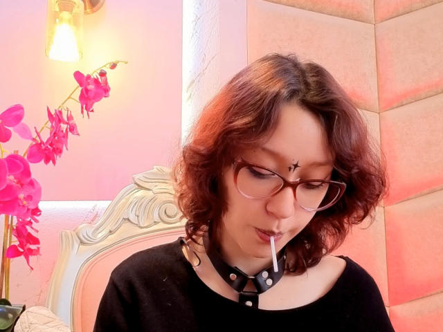 JinXkm with curly red-brown hair, glasses, black shirt, collar with studs, and cigarette, has a star tattoo on her forehead, pink orchids in the background.