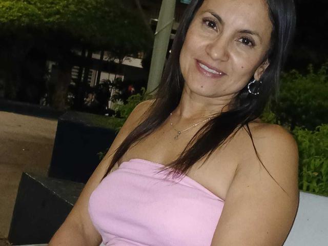 SolaraDubois with dark brown hair, pink strapless top, showing her tits, smiling, wearing hoop earrings, standing at night, greenery in the background.