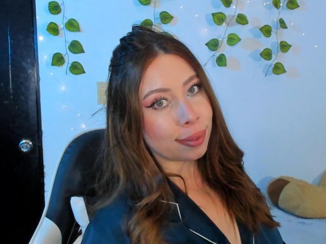 JennyLawry, avec ses cheveux bruns ondulés, son eye-liner rose et son rouge à lèvres, portant une robe noire, assise sur une chaise, regardant l'appareil photo avec un léger sourire..