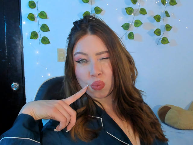 JennyLawry with pouty lips, winking, touching finger to mouth, wearing black robe with white stripes, brown wavy hair, light makeup, blue-lit background, leaf garland, and soft lights.
