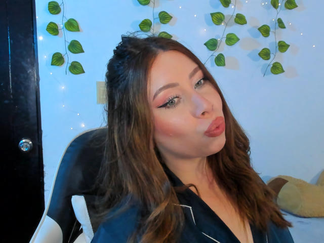 JennyLawry, pouty lips, brown wavy hair, black shirt, pink eyeshadow, chair, green leaf garland, blue wall, slightly tilted head, cute, sexy, soft lighting.