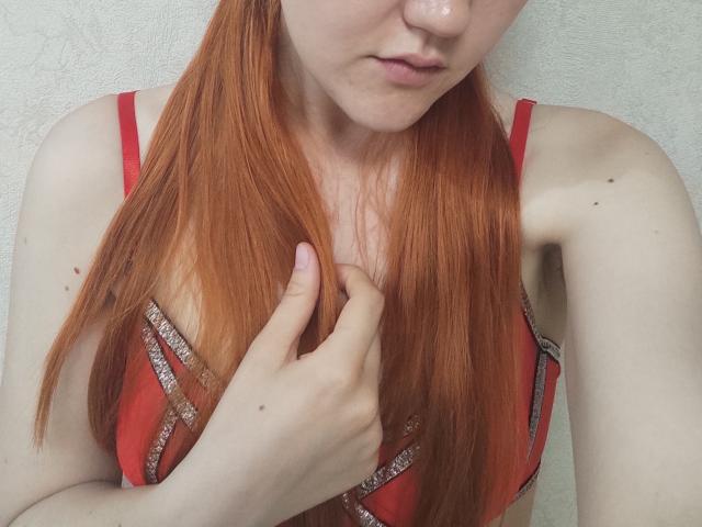SheritaChieng with red hair, pouty lips, fair skin, wearing a red bra with silver trim, hand in hair, showing small tits, and a few moles on her shoulder.