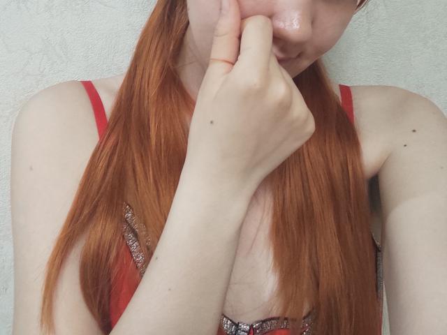 SheritaChieng with long red hair, red strap dress, pale skin, and hand over her mouth, showing her tits slightly. She has a small mole on her right arm.