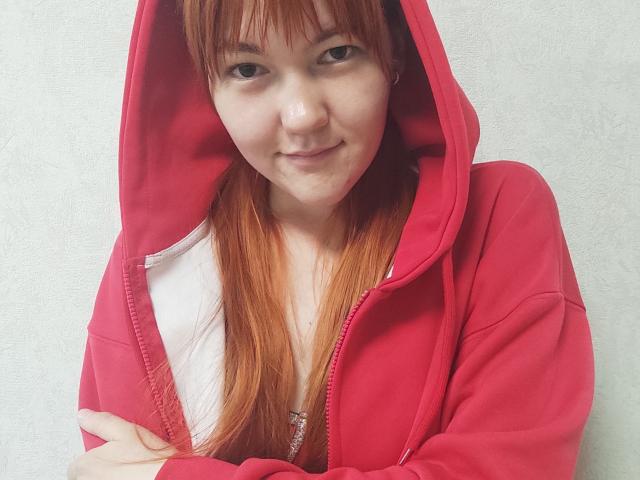 SheritaChieng has red hair, wearing a pink hoodie with the hood up, arms crossed, and a slight smile, showing her white shirt and necklace.