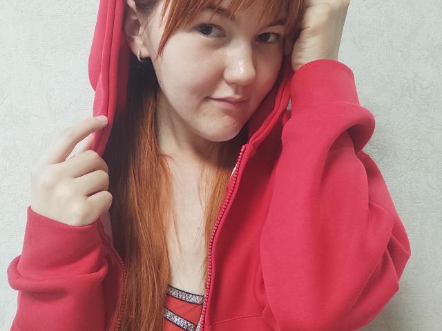 SheritaChieng, com cabelo ruivo, vestindo um moletom rosa, com as mãos dentro do capuz, um leve sorriso, sardas, um pequeno piercing no nariz, fundo de parede branca, close do tronco, casual, fofa..