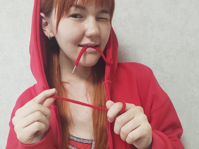 SheritaChieng, portant un sweat à capuche rouge, clignant de l'œil, mordant un fil rouge, le tenant avec ses mains, peau pâle, cheveux rouges lisses, petit nez, lèvres roses, mur blanc texturé en arrière-plan..