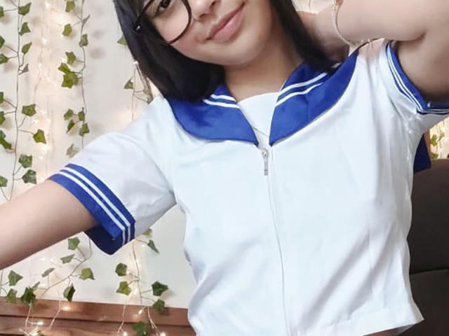 YayaLm with glasses, wearing a white and blue sailor-style top, smiling slightly, right arm raised, showing small tits, black hair, and fair skin, in a selfie.