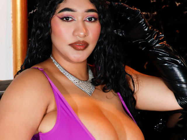 MelisaJackie with curly black hair, purple lipstick, and pink eyeshadow, wearing a low-cut purple top, black leather gloves, and a sparkling necklace, showing her large, shiny tits.