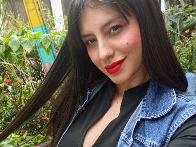 Madisson Fontaine, mit langen schwarzen Haaren, rotem Lippenstift und einer blauen Jeansjacke, trägt ein schwarzes Oberteil, hat ein süßes Lächeln und kleine Sommersprossen auf ihrer hellen Haut..