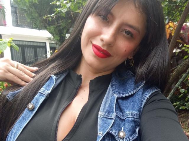 MadissonFontaine has long black hair, red lipstick, and is wearing a blue denim vest over a black top, with small tits visible. She's smiling and holding her hair.
