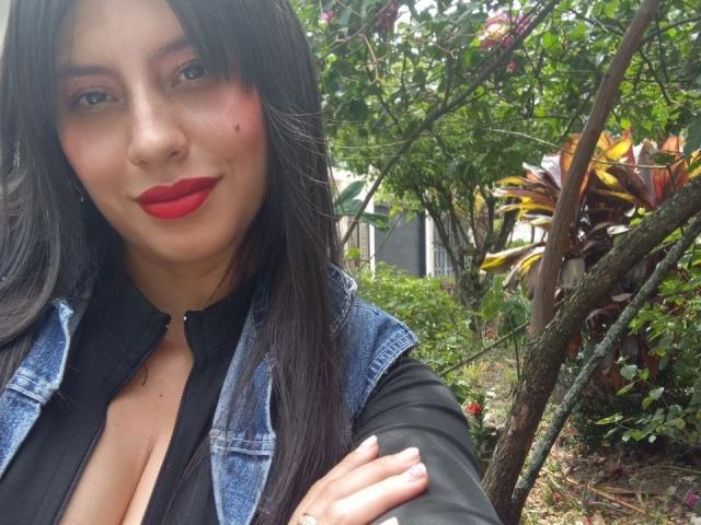 MadissonFontaine with red lipstick, black shirt, and blue denim vest, showing cleavage, standing in a garden with green leaves and trees. She has long black hair and a slight smile.