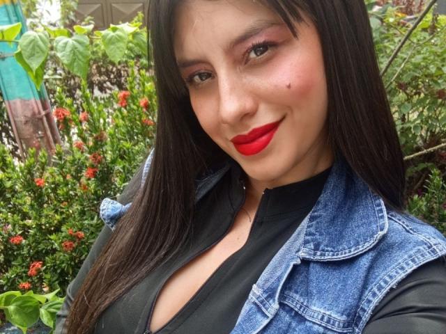 MadissonFontaine with red lips, black top, denim jacket, and long dark hair, showing a slight smile, with visible cleavage and a small mole on her right cheek.