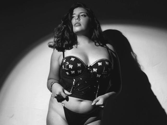 PaolaLyon with curvy body, wearing a dark, sequined, strapless top with star patterns, and black panties, standing in shadow, pulling down her top slightly, showing her belly.