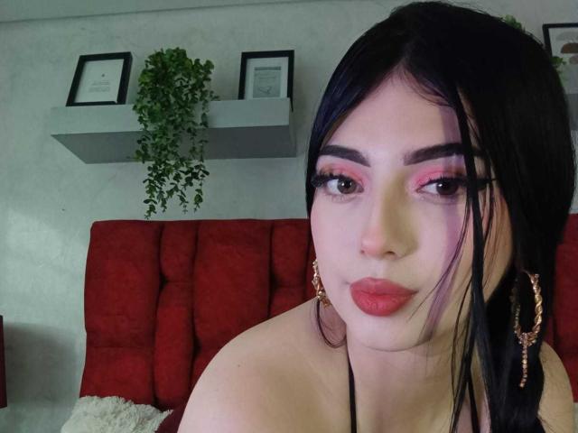 IsabellaaRossi has dark hair, pink eyeshadow, red lipstick, and black eyeliner. She's wearing a black halter top, with a red tufted headboard in the background.